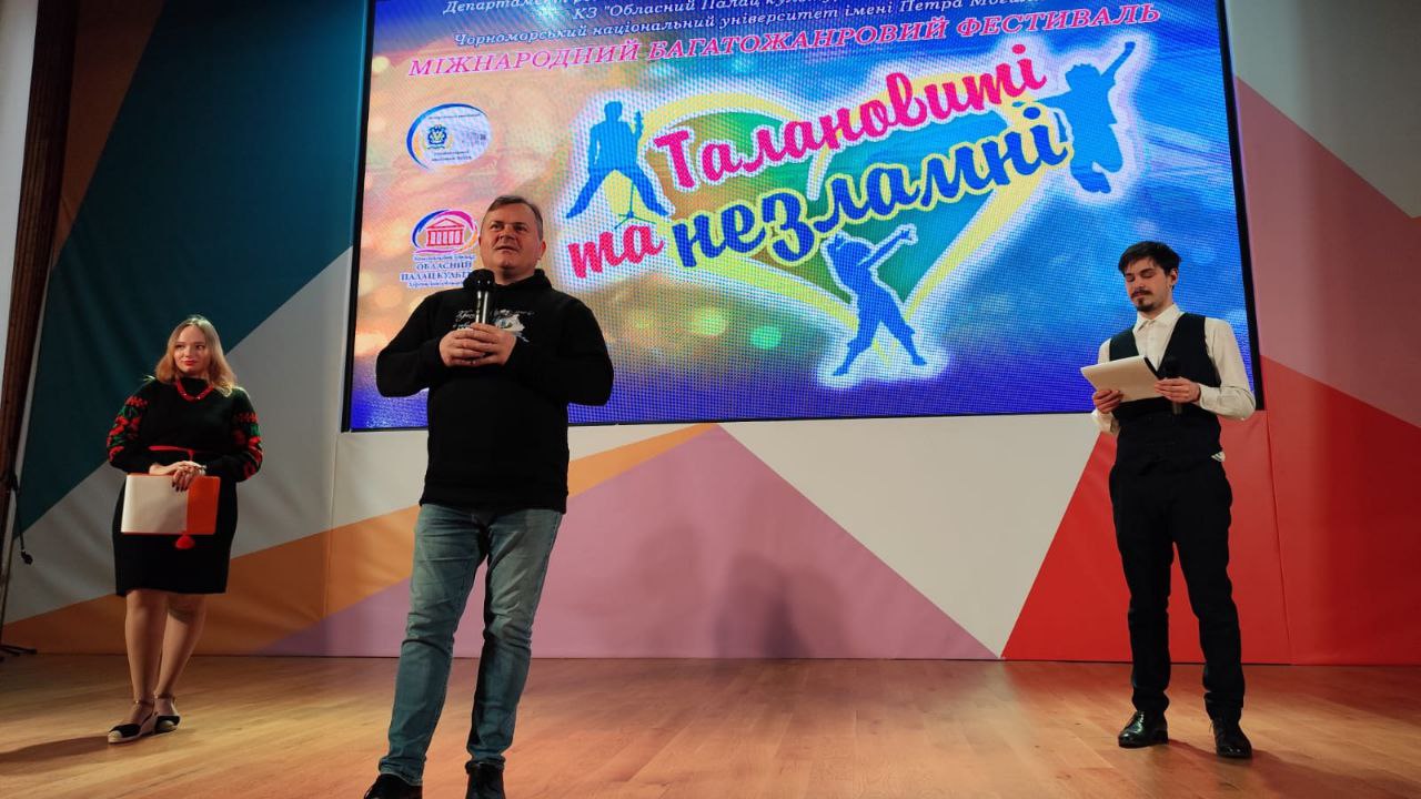 На сцені фестивалю стоять троє ведучих: чоловік у центрі говорить у мікрофон, ліворуч стоїть жінка у вишиванці, а праворуч — молодий чоловік із нотатками. На задньому плані — великий екран із логотипом фестивалю «Талановиті та незламні».