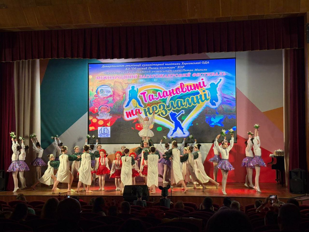 На сцені актового залу виступає великий дитячий танцювальний колектив у різнобарвних сценічних костюмах. На задньому плані розташовано великий екран із написом «Міжнародний багатожанровий фестиваль „Талановиті та незламні“», а в залі присутні глядачі.