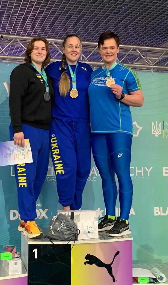 Три спортсменки у синьо-жовтій формі збірної України стоять на п'єдесталі пошани. Вони тримають медалі та диплом на фоні брендованої стіни зі спортивною символікою.