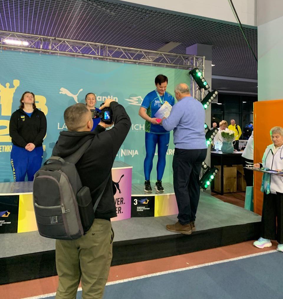 Процес нагородження на спортивних змаганнях: офіційна особа вручає диплом спортсменці, яка стоїть на третій сходинці п'єдесталу. На передньому плані фотограф знімає подію на камеру, а на фоні розміщено банер із логотипами спортивних організацій.