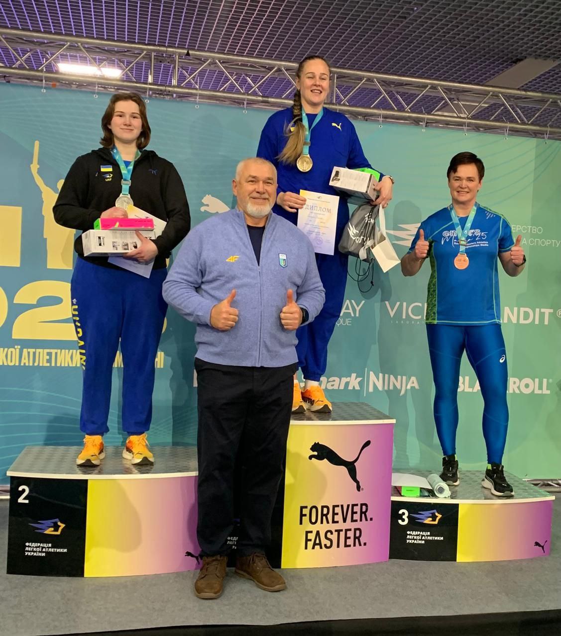 Нагородження переможців спортивних змагань: три спортсменки у формі збірної України з медалями та дипломами стоять на п'єдесталі. Попереду них стоїть тренер, який показує жест «великий палець вгору» на фоні банера Федерації легкої атлетики України.