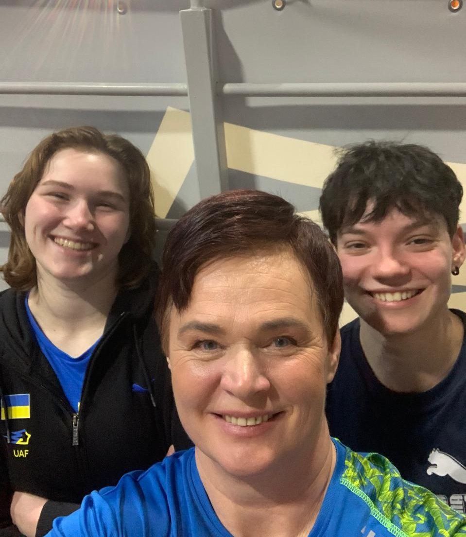 Троє усміхнених людей роблять спільне селфі у спортивному залі на фоні шведської стінки. Одна з учасниць одягнена у синьо-жовту спортивну форму з логотипом «UAF» (Федерація легкої атлетики України).