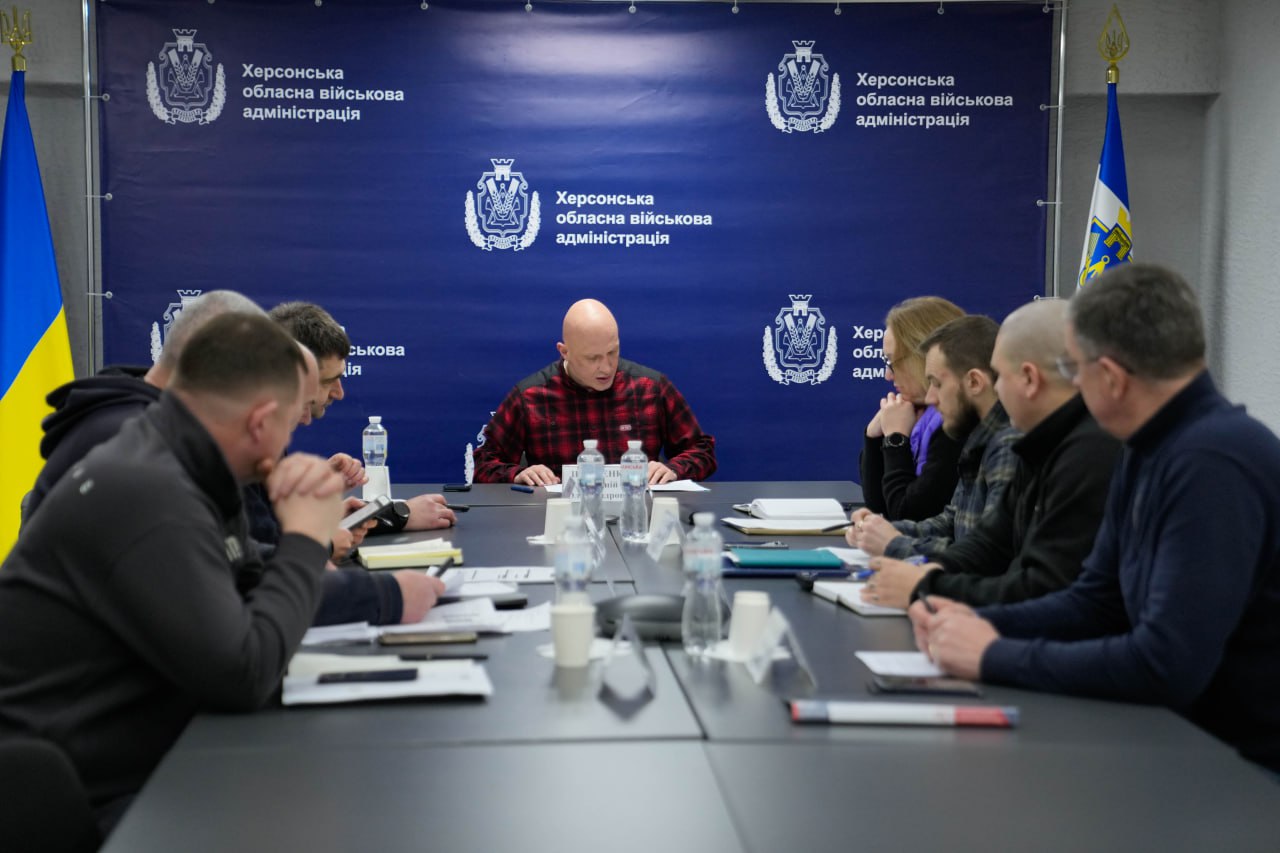 Група людей на офіційній нараді за довгим столом. У центрі — чоловік у червоно-чорній сорочці в клітинку. На фоні — синій брендований банер Херсонської ОВА та державні прапори.