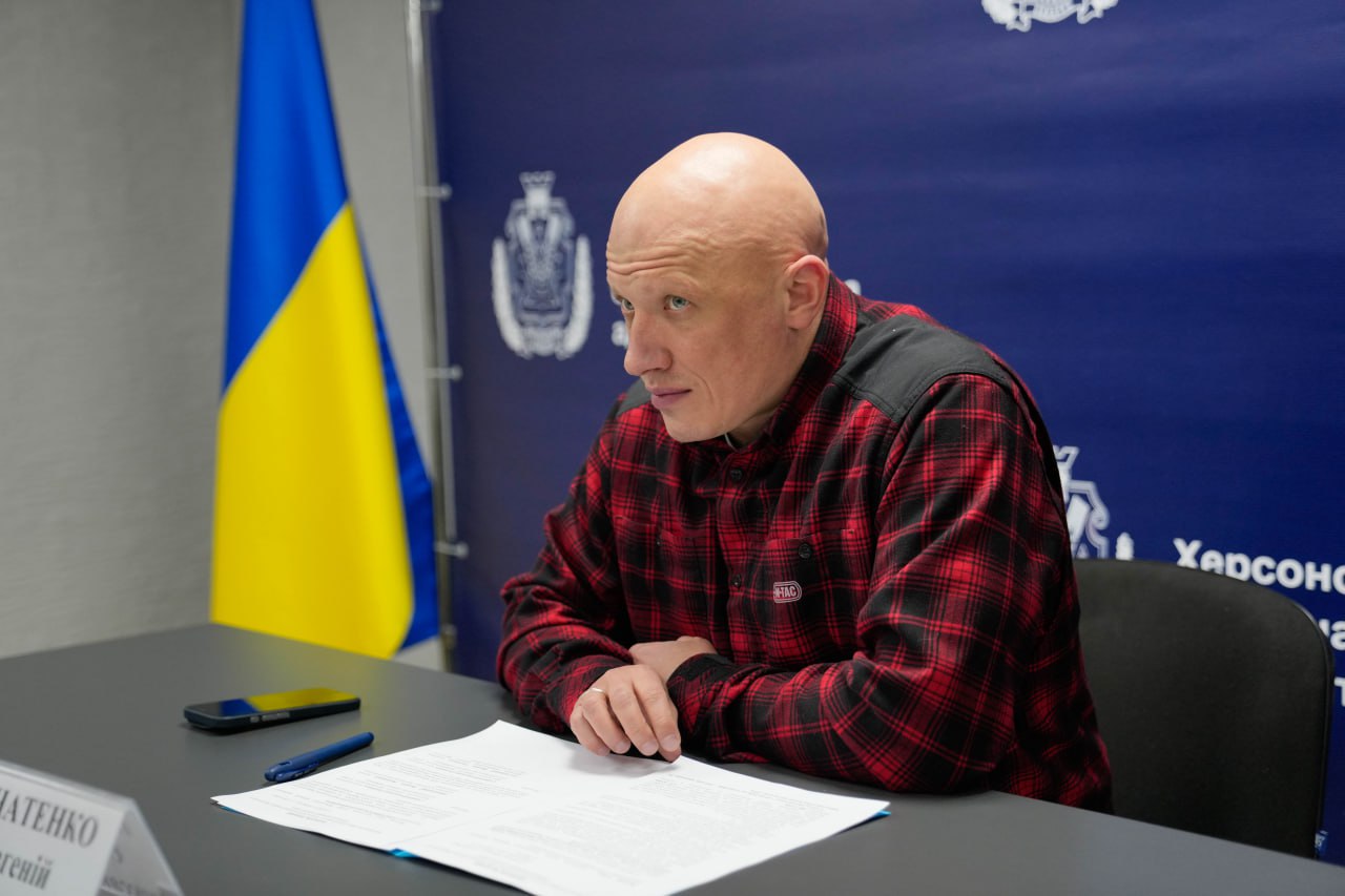 Чоловік у сорочці в клітинку за робочим столом із документами на фоні прапора України та синього банера з офіційною символікою.