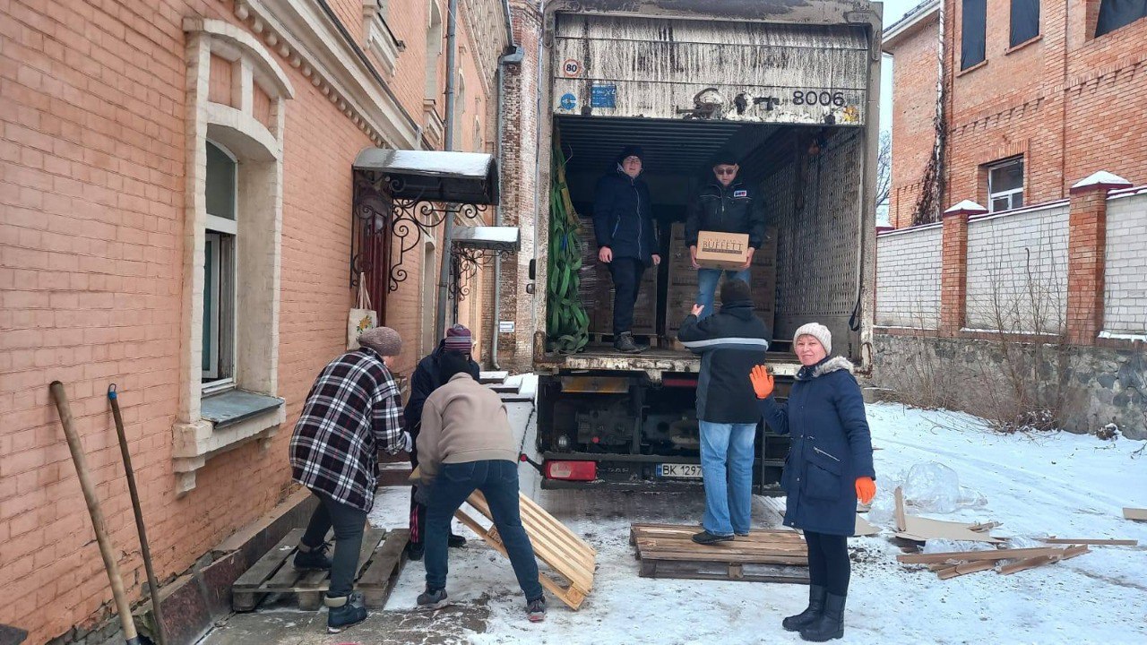 Група людей розвантажує коробки з вантажівки на засніженому подвір'ї біля цегляної будівлі. Двоє чоловіків подають вантаж із фури, інші приймають його на землі, використовуючи дерев'яні піддони.