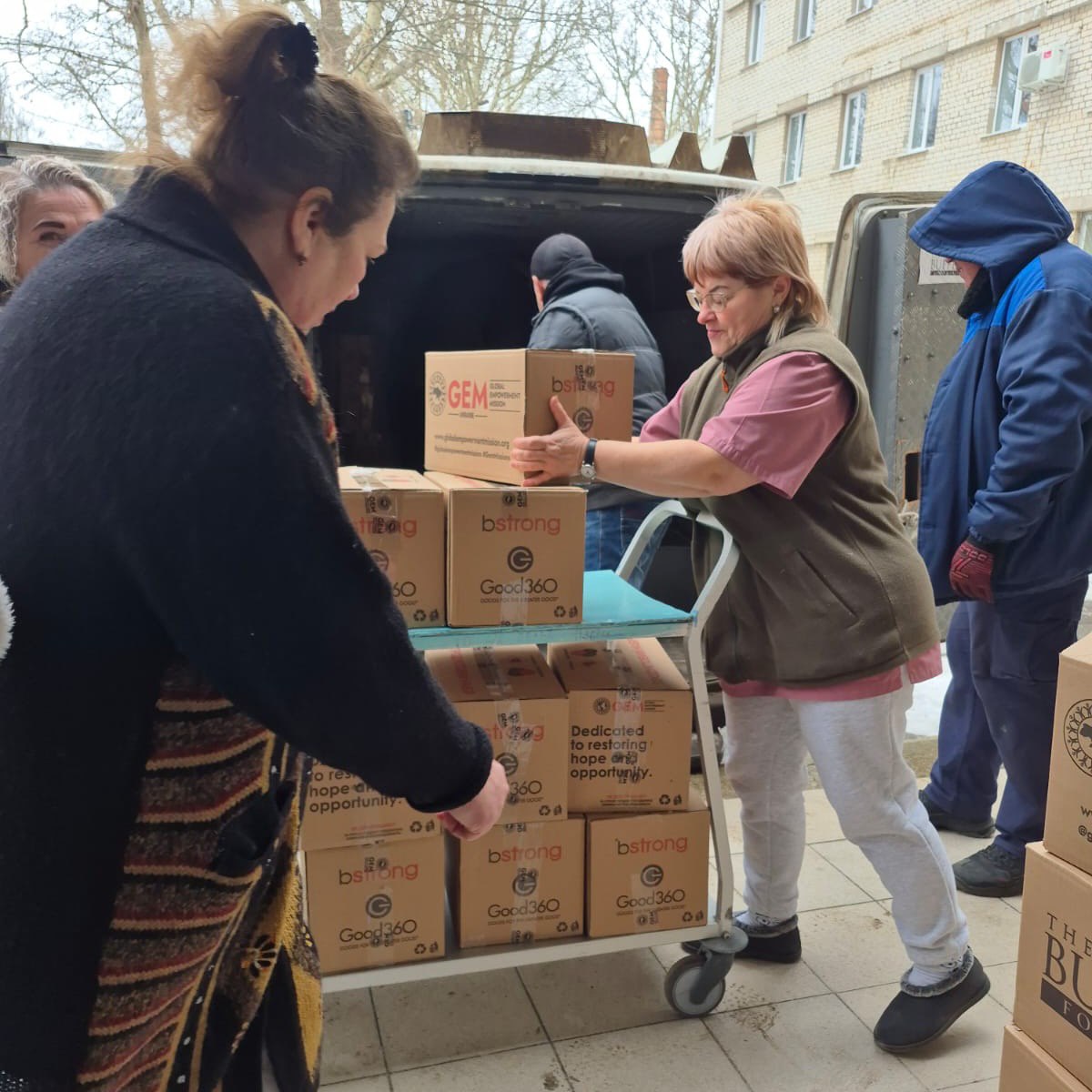 Люди розвантажують гуманітарну допомогу з мікроавтобуса. Жінка складає коробки з маркуванням благодійних організацій на вантажний візок, поруч інші допомагають із розвантаженням.