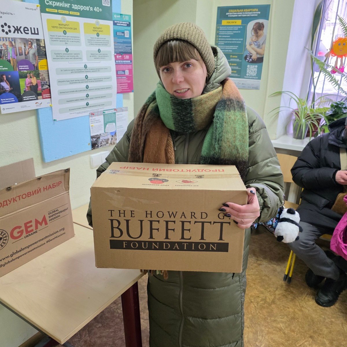 Жінка в теплому верхньому одязі тримає велику картонну коробку з гуманітарною допомогою. На коробці нанесено логотип благодійної організації. Вона стоїть у приміщенні поруч із інформаційним стендом та іншими коробками.