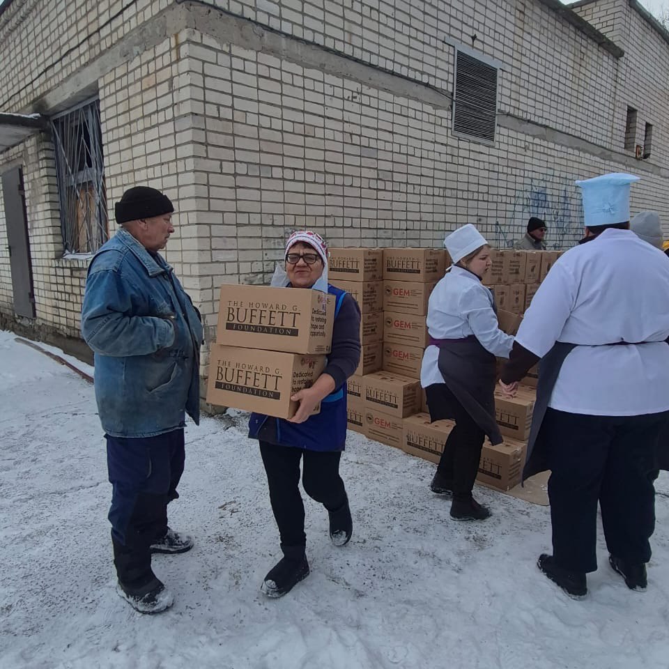 Жінка несе дві коробки з гуманітарною допомогою на тлі цегляної будівлі. Поруч стоять інші люди, зокрема працівники у білій кухонній формі, які складають коробки у штабелі на засніженому подвір'ї.