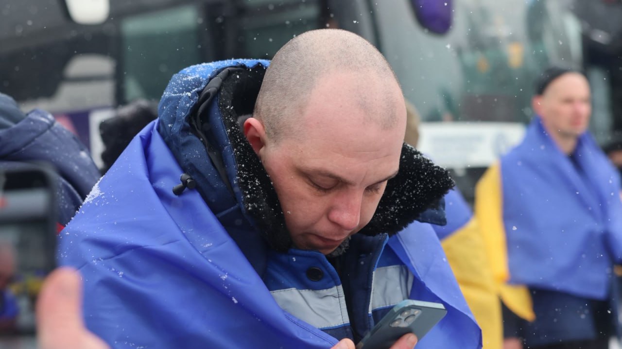 Чоловік, загорнутий у синьо-жовтий прапор, зосереджено дивиться у свій смартфон. Він стоїть на вулиці під час снігопаду на фоні великого автобуса та інших людей у зимовому одязі.