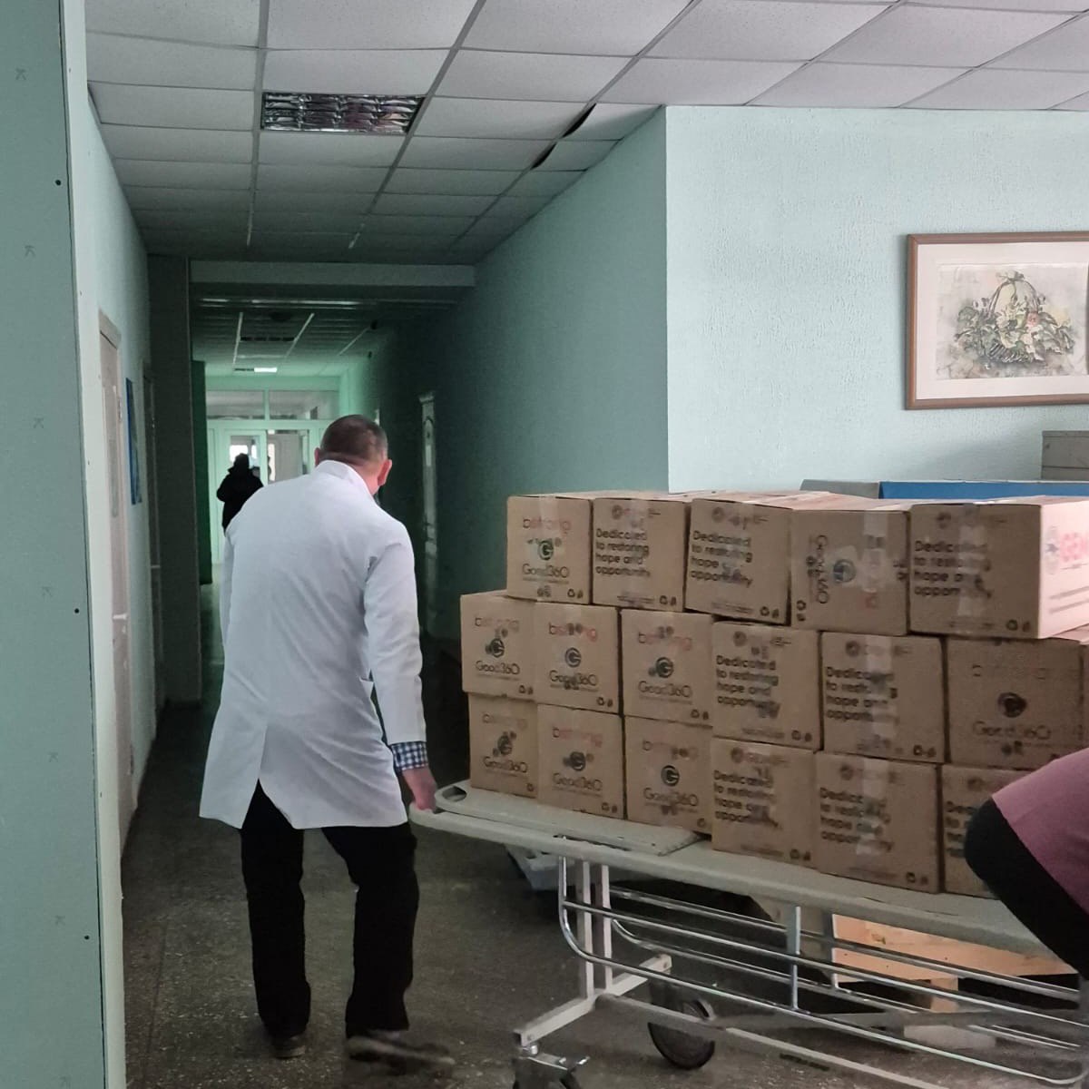 Чоловік у білому медичному халаті везе коридором закладу вантажну каталку, вщент заповнену коробками з гуманітарною допомогою. На коробках видно маркування благодійних організацій.