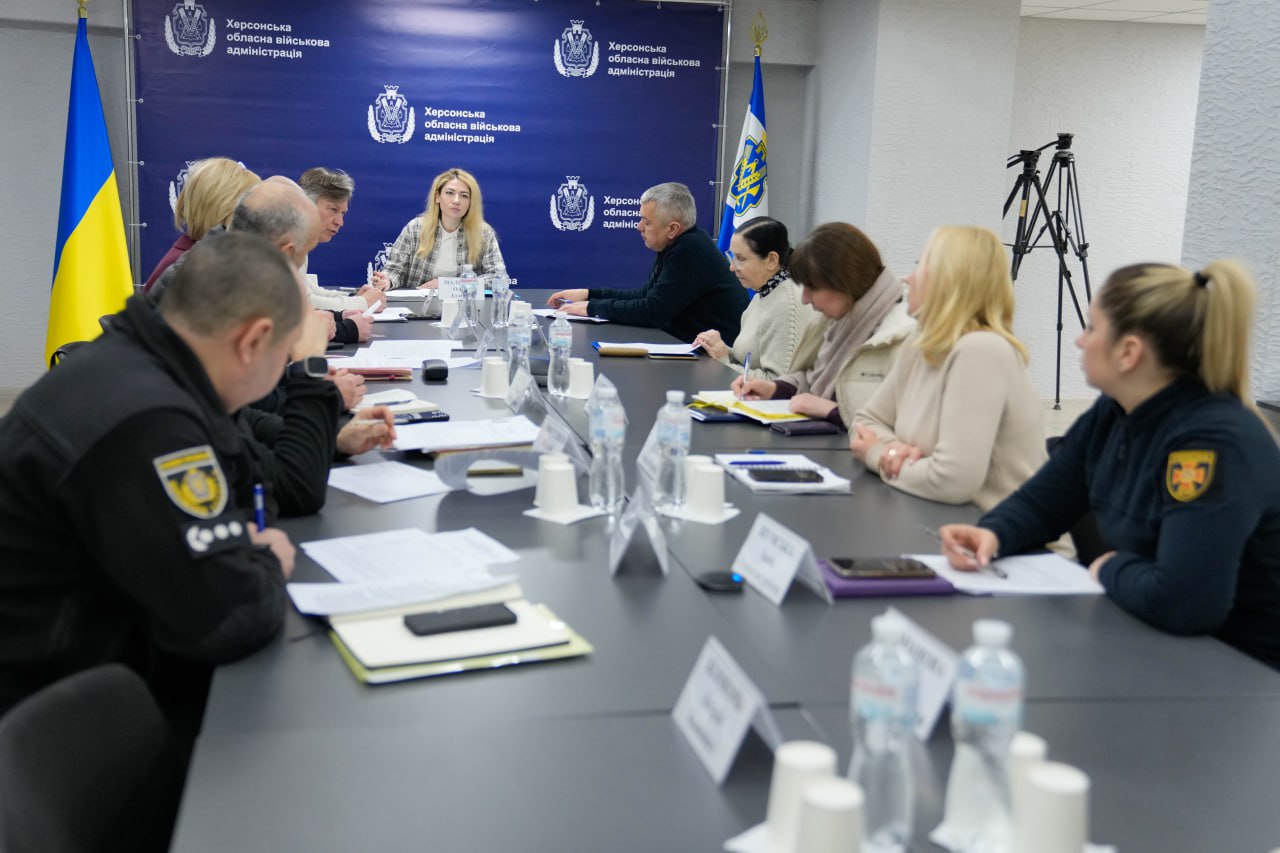 Група людей сидить за великим столом під час наради в приміщенні з прапорами України та банером Херсонської обласної військової адміністрації. Учасники обговорюють документи, занотовують інформацію, а в залі встановлено штатив для відеозйомки.