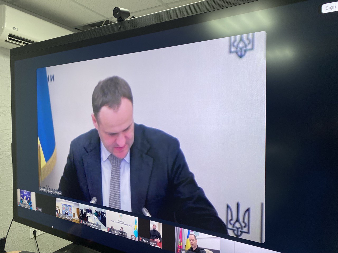 Екран монітора, на якому транслюється онлайн-нарада за участю посадовців. У центрі — основне вікно з доповідачем на фоні державного прапора та герба України, а внизу — іконки інших учасників відеоконференції від різних обласних адміністрацій.