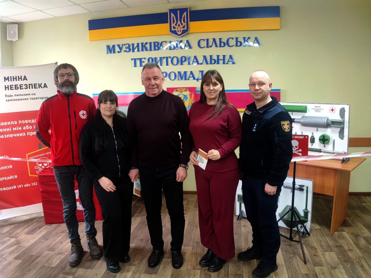 П’ятеро людей стоять у приміщенні на фоні стіни з написом «Музиківська сільська територіальна громада» та державною символікою України. Серед присутніх — представник Червоного Хреста та рятувальник ДСНС. Поруч розміщено інформаційні стенди та макети, присвячені мінній безпеці.
