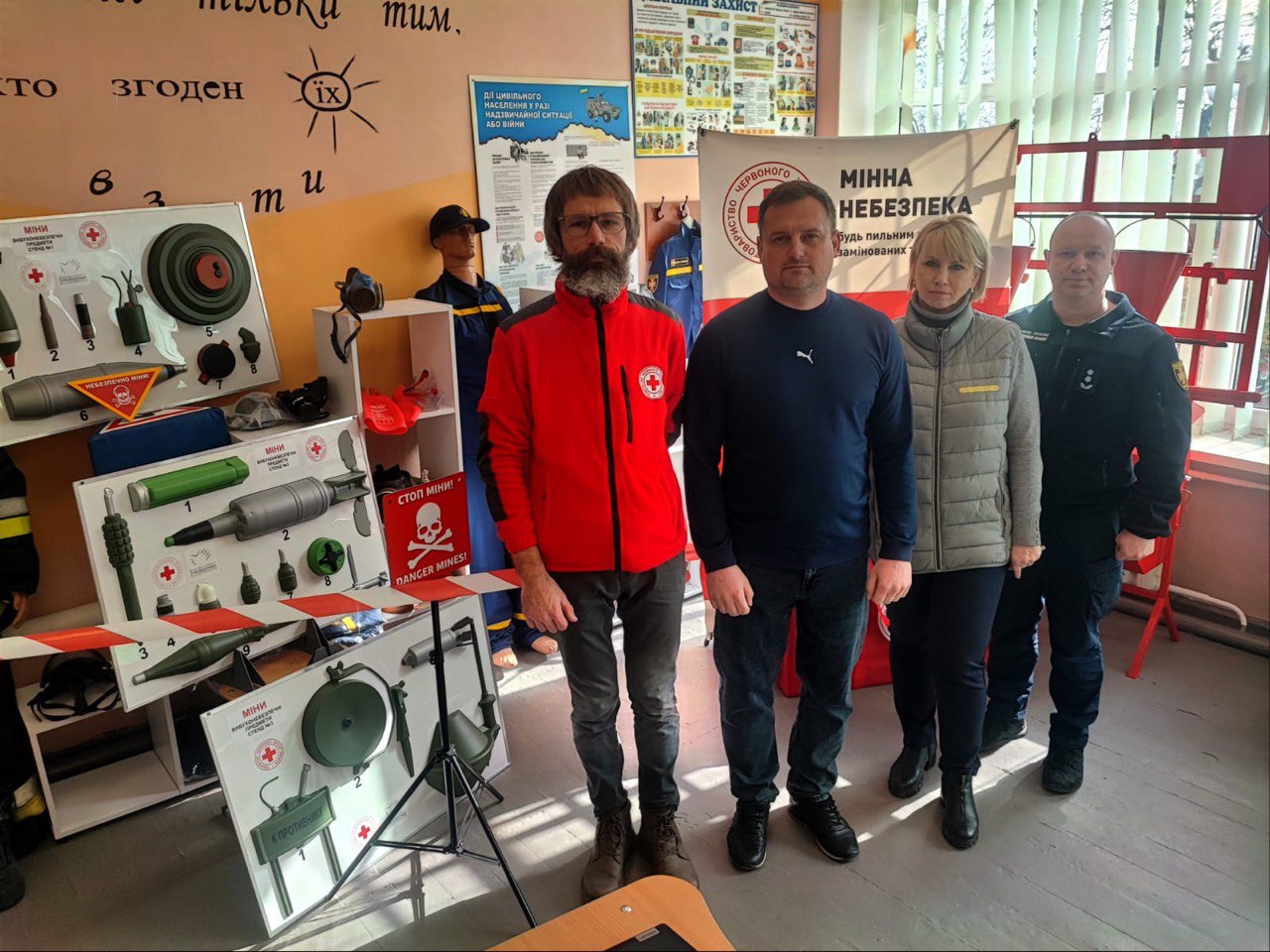 Четверо людей стоять у спеціально обладнаному класі безпеки. На задньому плані та поруч із ними розміщено численні інформаційні стенди, макети мін та снарядів, а також банер Товариства Червоного Хреста України з написом «Мінна небезпека». Серед групи — фахівець ДСНС та волонтер Червоного Хреста.