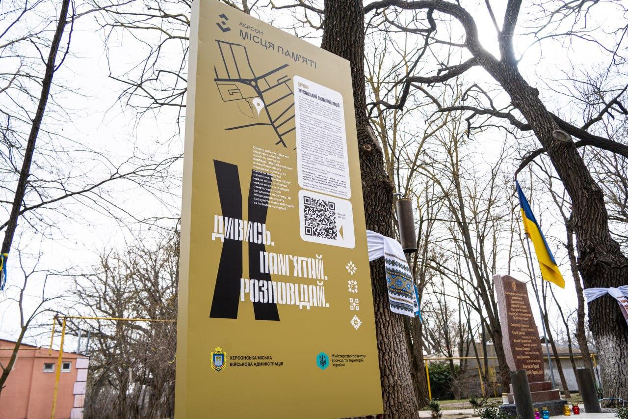 На передньому плані встановлено високу інформаційну панель жовтого кольору з написом «Херсон. Місця пам'яті». На задньому плані видно дерева, прапор України та меморіальну плиту.