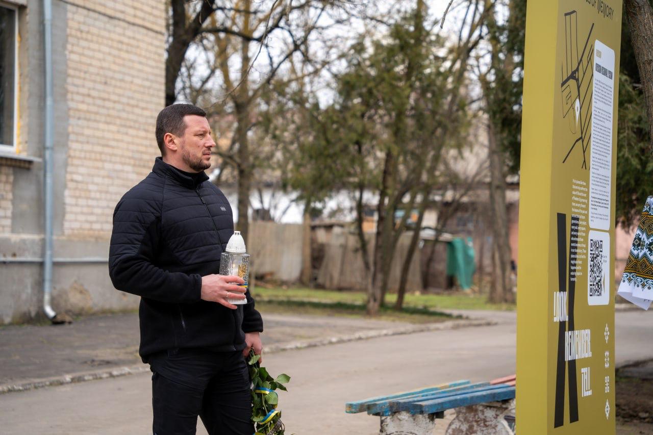 Чоловік у чорному одязі стоїть на вулиці поруч із меморіальною стелою, тримаючи в руках лампадку та квіти з синьо-жовтою стрічкою.