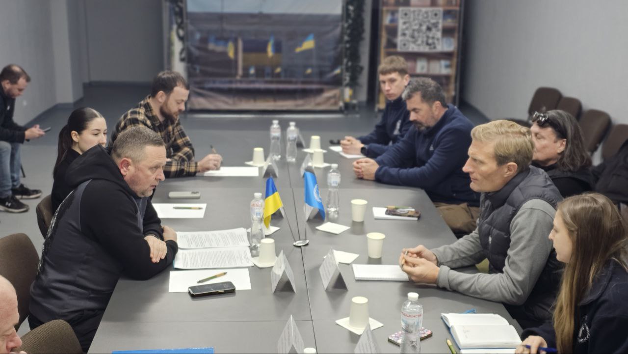 Група людей сидить за довгим столом під час ділової зустрічі або переговорів. На столі встановлені маленькі прапори України та Організації Об'єднаних Націй, лежать документи, блокноти та пляшки з водою. Учасники зосереджено спілкуються в офіційній робочій атмосфері.