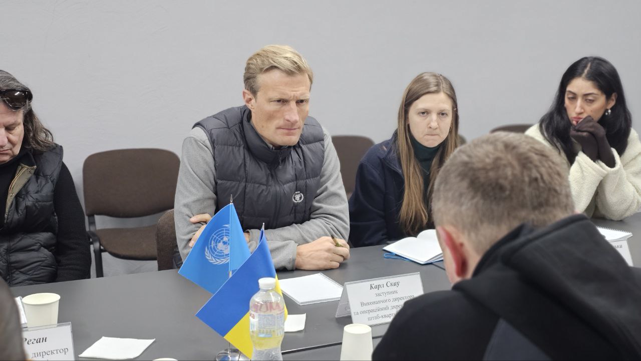 Представники делегацій за столом переговорів. У фокусі — учасники, що ведуть діалог, на передньому плані стоять настільні прапорці України та ООН поруч із іменною табличкою.