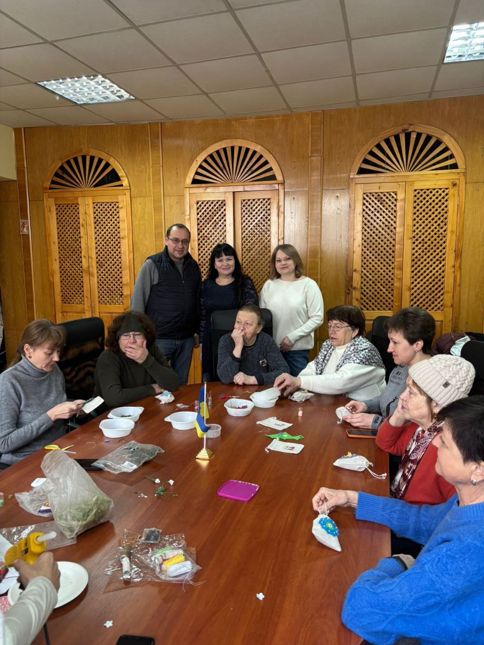 Група людей за великим столом під час творчого заняття в приміщенні з дерев'яним оздобленням стін. Учасники виготовляють дрібні сувеніри, використовуючи нитки, тканину та підручні матеріали. У центрі столу стоїть маленький прапор України.