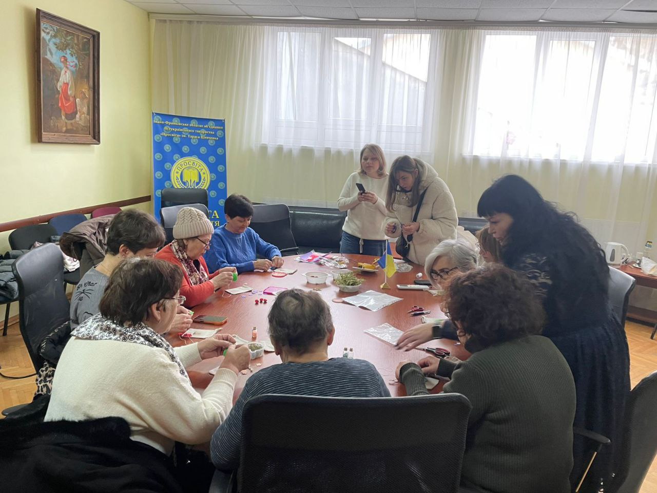 Група жінок за круглим столом займається рукоділлям у світлій кімнаті. Вони зосереджено працюють над дрібними виробами, використовуючи нитки та тканину. На фоні видно вікна з тюлем, інформаційний банер та картину на стіні.