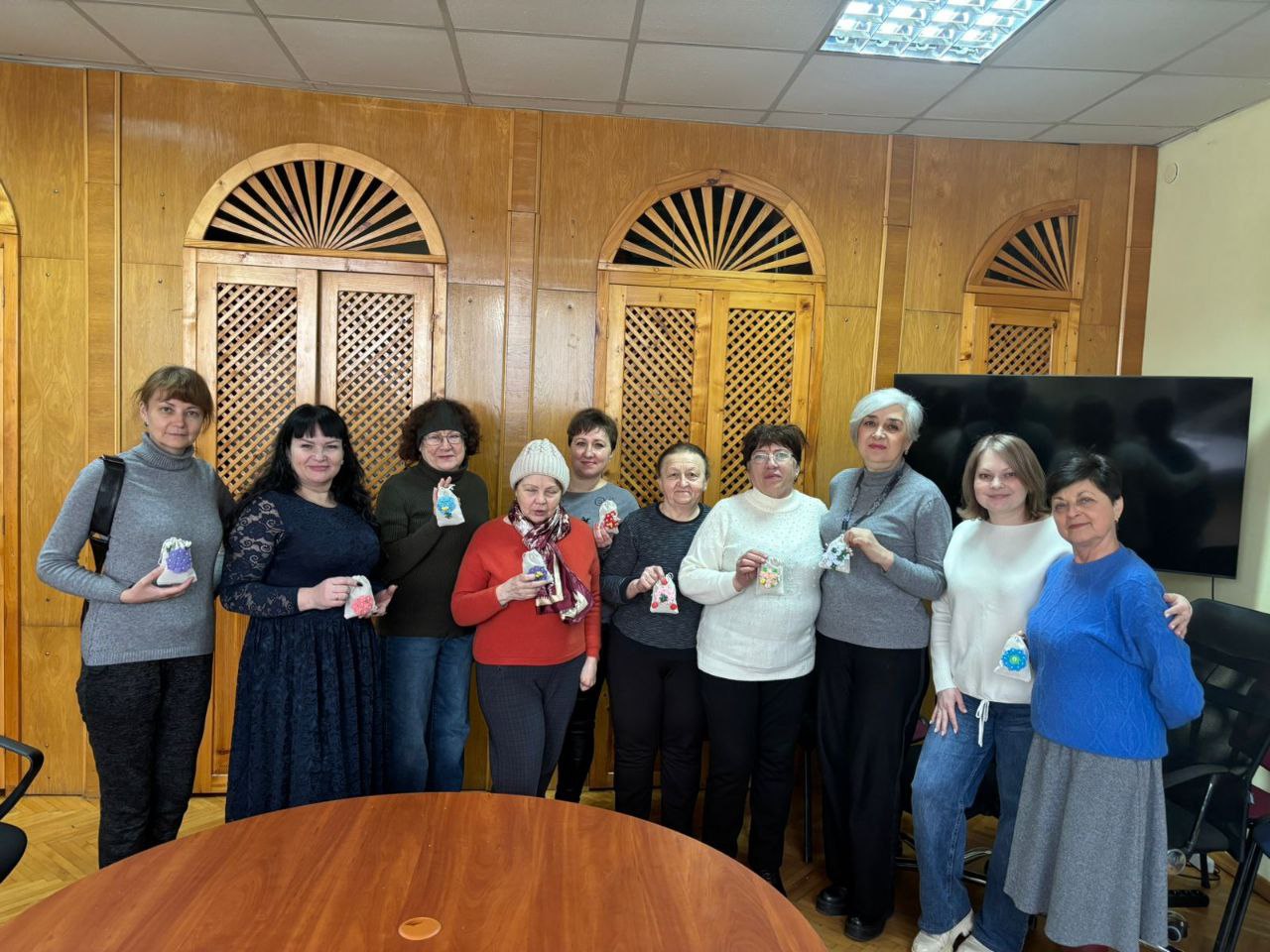 Ответ Gemini Група жінок стоїть у світлій кімнаті з дерев’яним оздобленням стін. Кожна тримає в руках невеликий вишитий або декорований мішечок ручної роботи. Учасниці позують для спільного фото поруч із круглим столом.