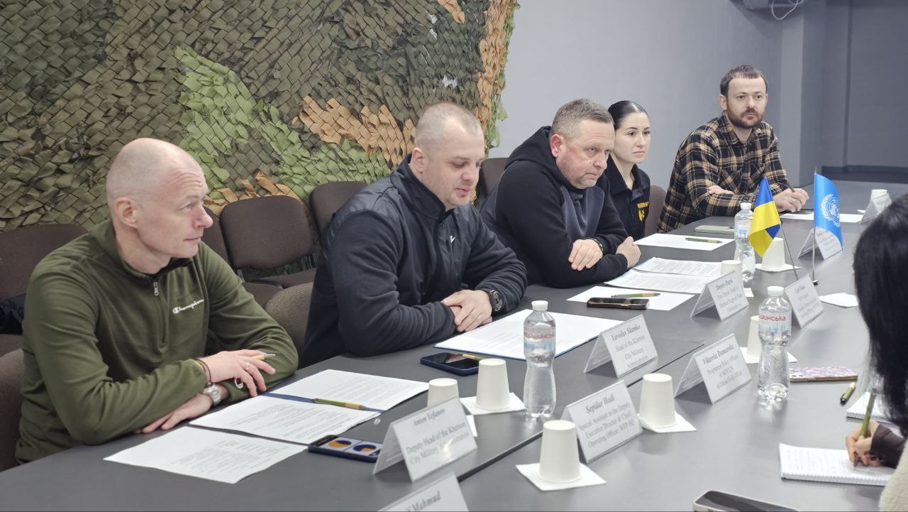 Учасники делегації сидять за столом під час офіційних перемовин. Перед ними лежать роздруковані документи та таблички з іменами. На фоні видно маскувальну сітку, що закриває частину стіни. На столі присутня символіка України та ООН.