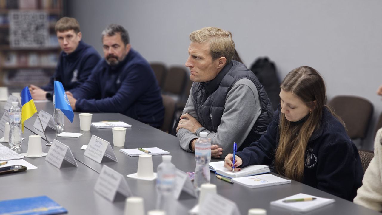 Учасники зустрічі сидять у ряд, уважно слухаючи доповідача. Одна з учасниць робить записи в блокноті. На столі видно символіку України та ООН, а також паперові стаканчики та канцелярське приладдя.