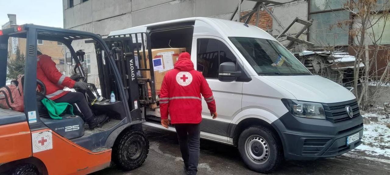 Навантажувач переміщує велику коробку з маркуванням гуманітарної допомоги в білий вантажний мікроавтобус. Працівник у червоному спецодязі з логотипом організації координує процес завантаження на відкритому майданчику.