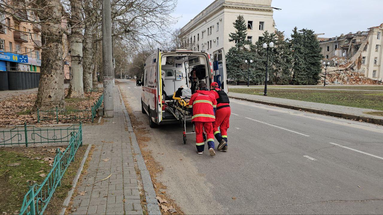 Медики завантажують пацієнта на ношах до автомобіля швидкої допомоги на міській вулиці. На задньому плані видно пошкоджену обстрілами багатоповерхову будівлю та порожній тротуар з деревами.