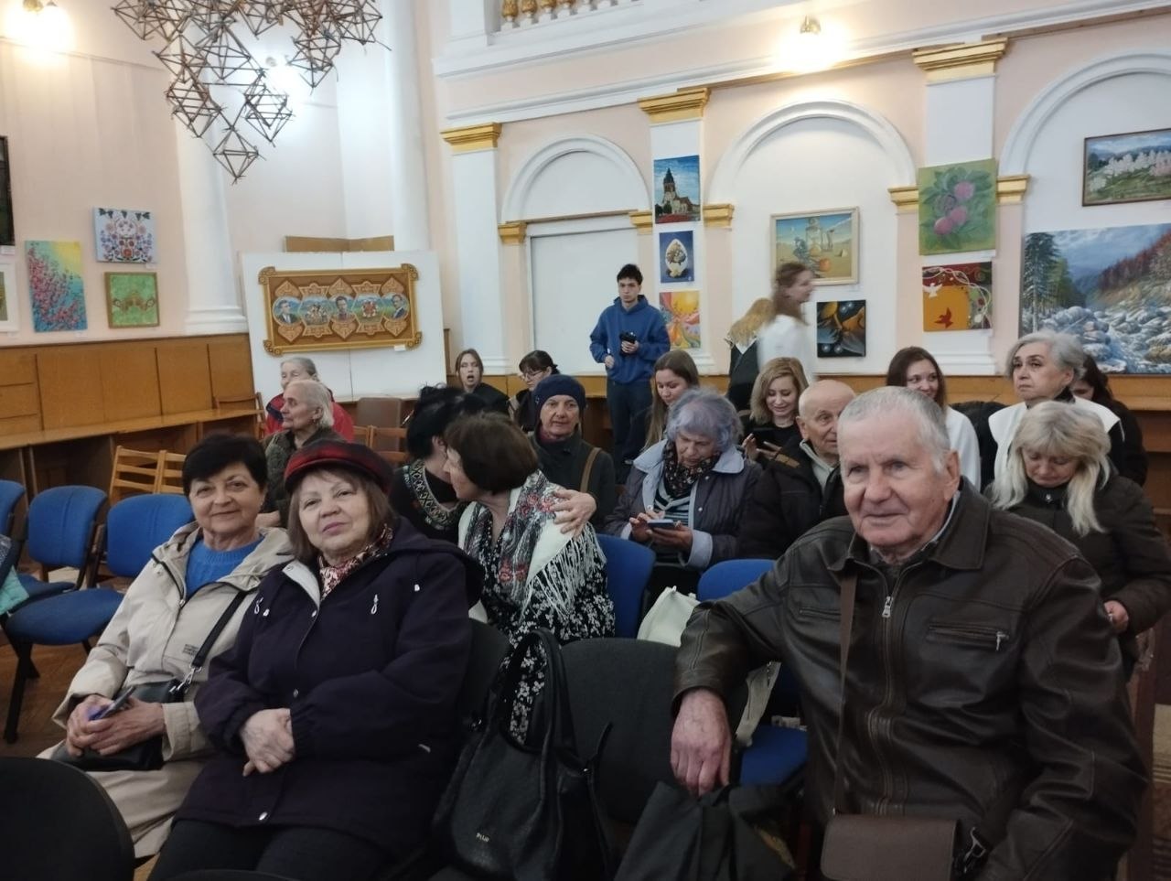 Група людей різного віку сидить у світлій залі на фоні художньої виставки. На стінах розвішані картини, а вгорі видно декоративну підвісну конструкцію.