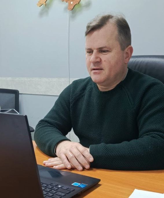 Чоловік у темно-зеленому светрі сидить за робочим столом перед відкритим ноутбуком. На задньому плані видно частину стіни з декоративною мапою України.