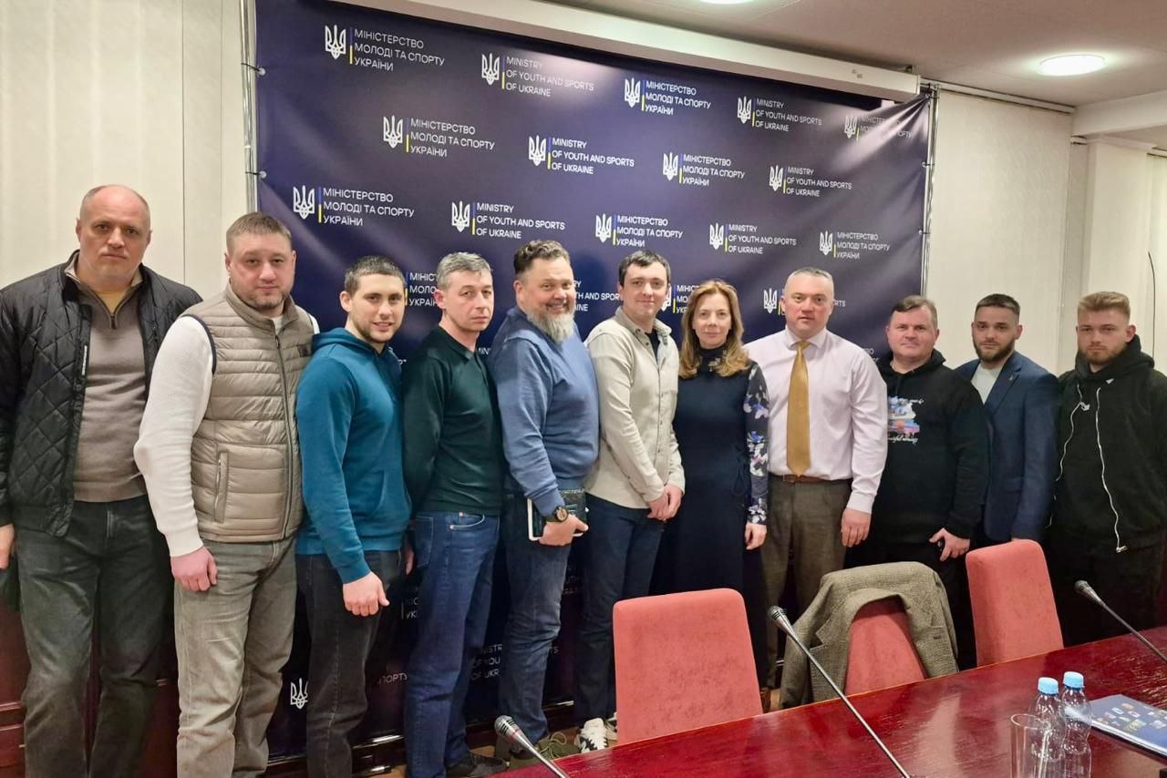 Групова фотографія одинадцяти осіб у приміщенні на фоні синього брендованого банера Міністерства молоді та спорту України. Учасники зустрічі стоять у ряд, вдягнені у діловий та повсякденний одяг. На передньому плані видно частину столу для засідань із документами та водою.