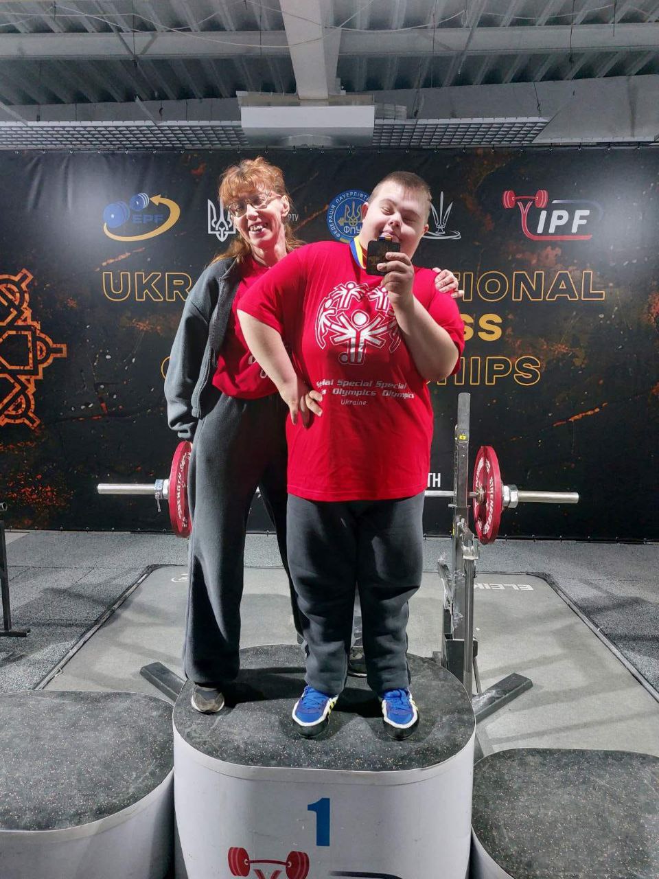 Спортсмен у червоній футболці Спеціальної Олімпіади стоїть на першій сходинці п'єдесталу, піднявши медаль до обличчя. Позаду нього стоїть жінка, яка його підтримує. Вони позують на фоні професійного спортивного обладнання.