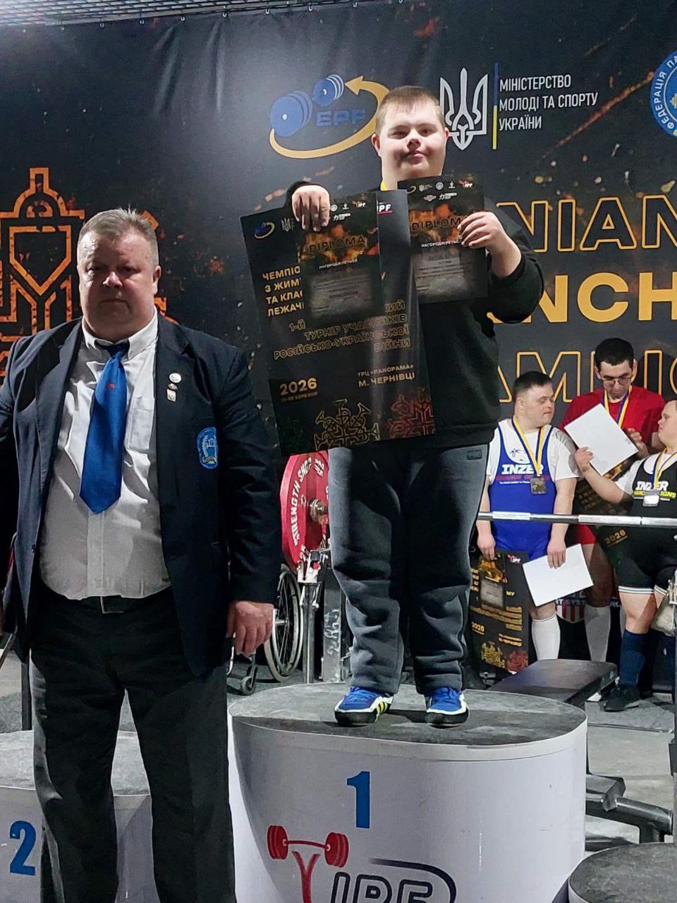 Молодий спортсмен стоїть на першій сходинці п'єдесталу пошани, тримаючи в руках дипломи та плакат змагань з пауерліфтингу. Поруч стоїть офіційний представник у діловому костюмі. На фоні — банер із логотипом Міністерства молоді та спорту України та інша символіка чемпіонату.