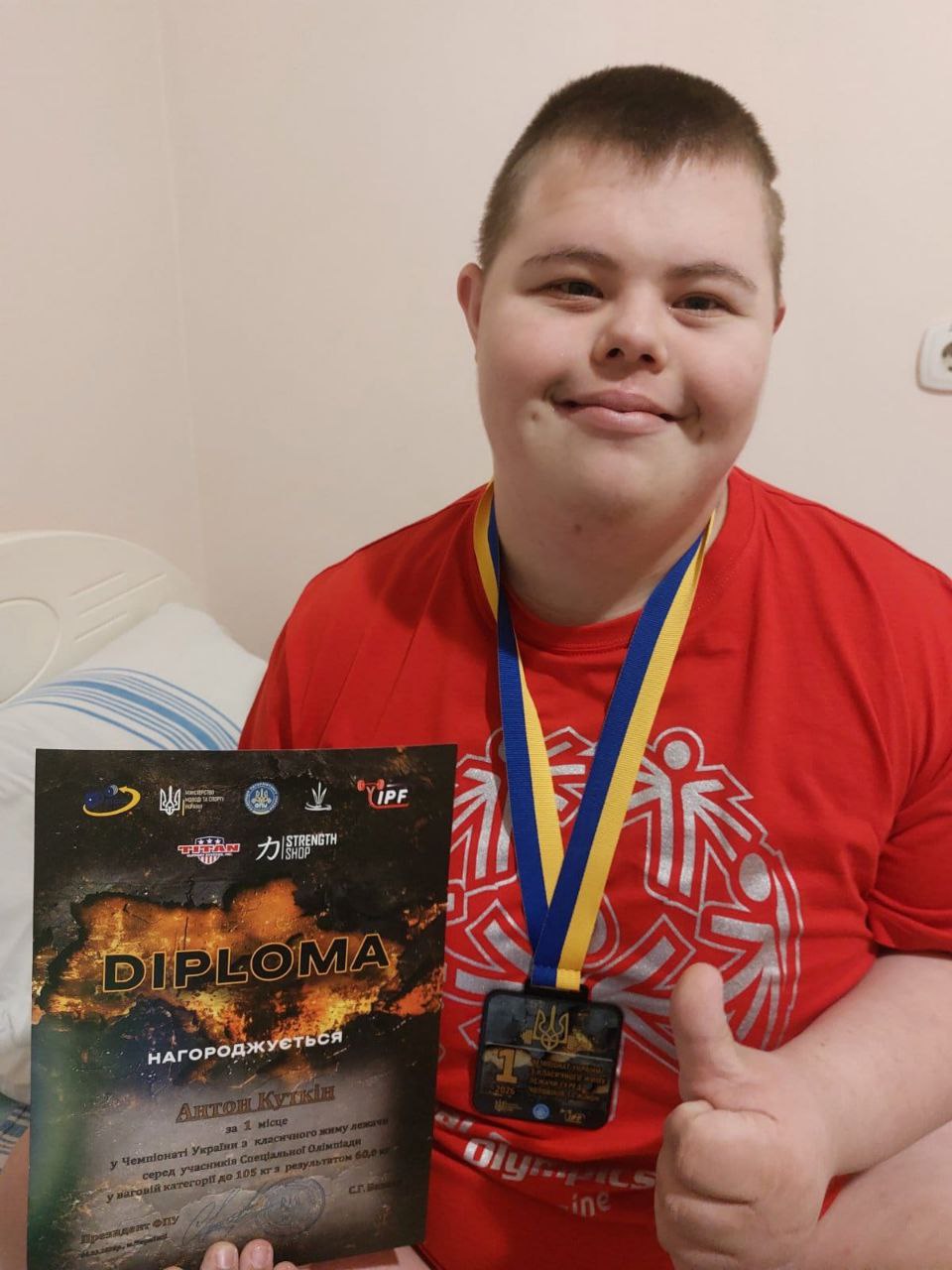 Учасник змагань у червоній футболці з медаллю на грудях тримає диплом за перше місце у чемпіонаті України з класичного жиму лежачи серед учасників Спеціальної Олімпіади та показує жест «великий палець вгору».
