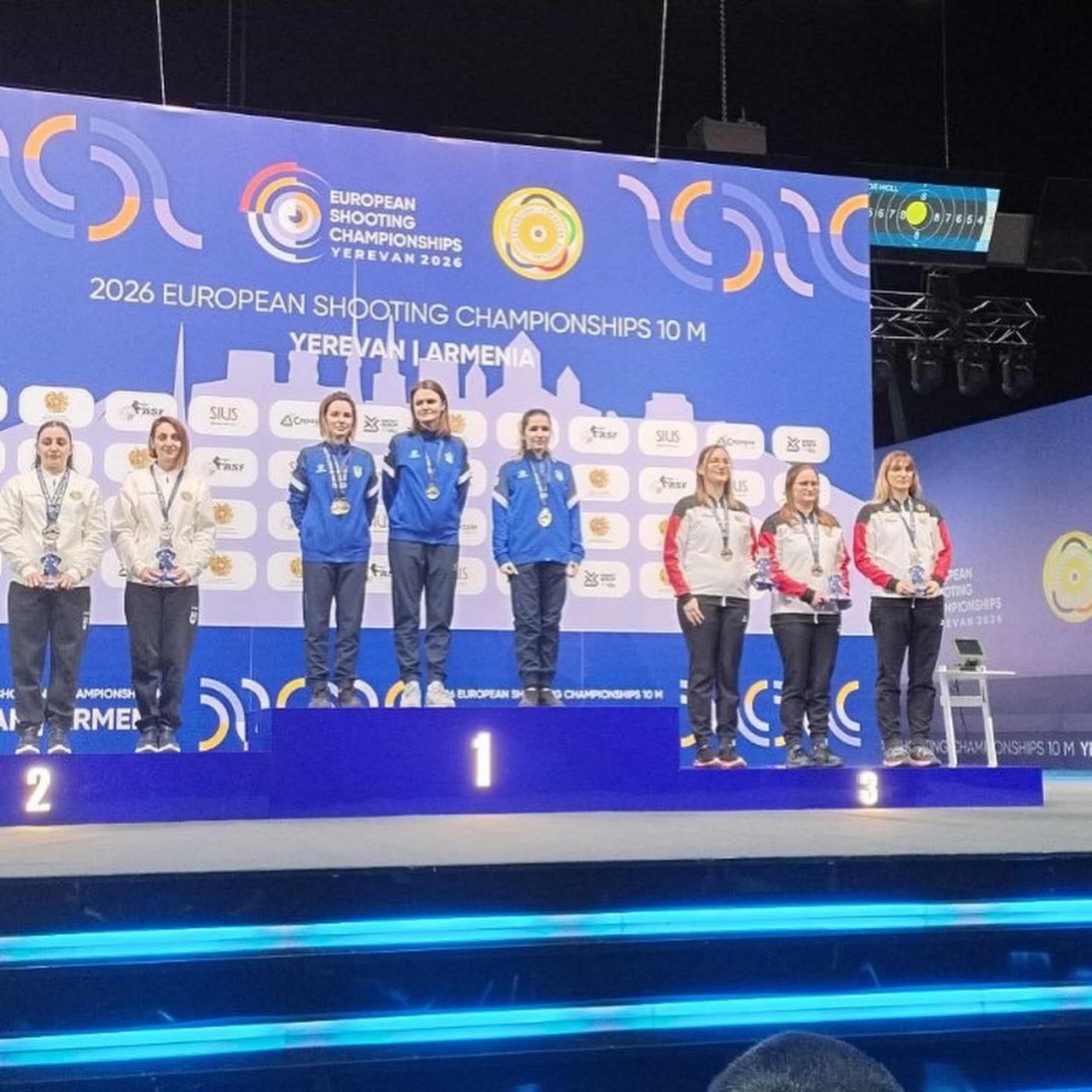 Вісім спортсменок стоять на п'єдесталі пошани під час церемонії нагородження на Чемпіонаті Європи зі стрільби 2026 року в Єревані. Учасниці на першому місці одягнені в синьо-жовту спортивну форму. На великому екрані позаду них розміщено логотип змагань та силуети міста.