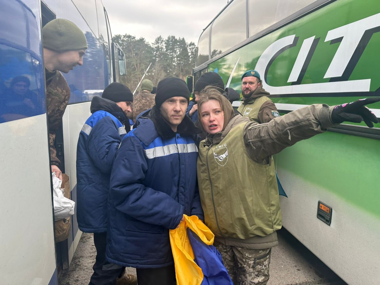 Група людей стоїть між двома великими автобусами просто неба. У центрі — чоловік у синій куртці, який тримає в руках синьо-жовтий прапор. Поруч із ним жінка у військовій формі вказує рукою вбік. Навколо знаходяться інші особи у форменому та цивільному одязі на фоні лісу.