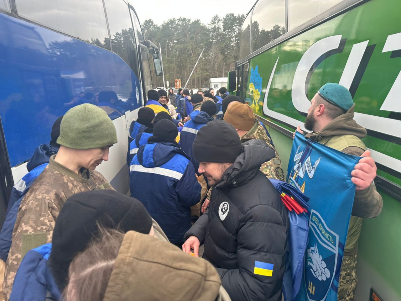 Натовп людей у військовому та цивільному одязі зібрався у вузькому проході між двома великими автобусами. Праворуч чоловік тримає велике синє полотно з емблемою та написом. На бічній панелі зеленого автобуса видно наклейку з картою України в національних кольорах.