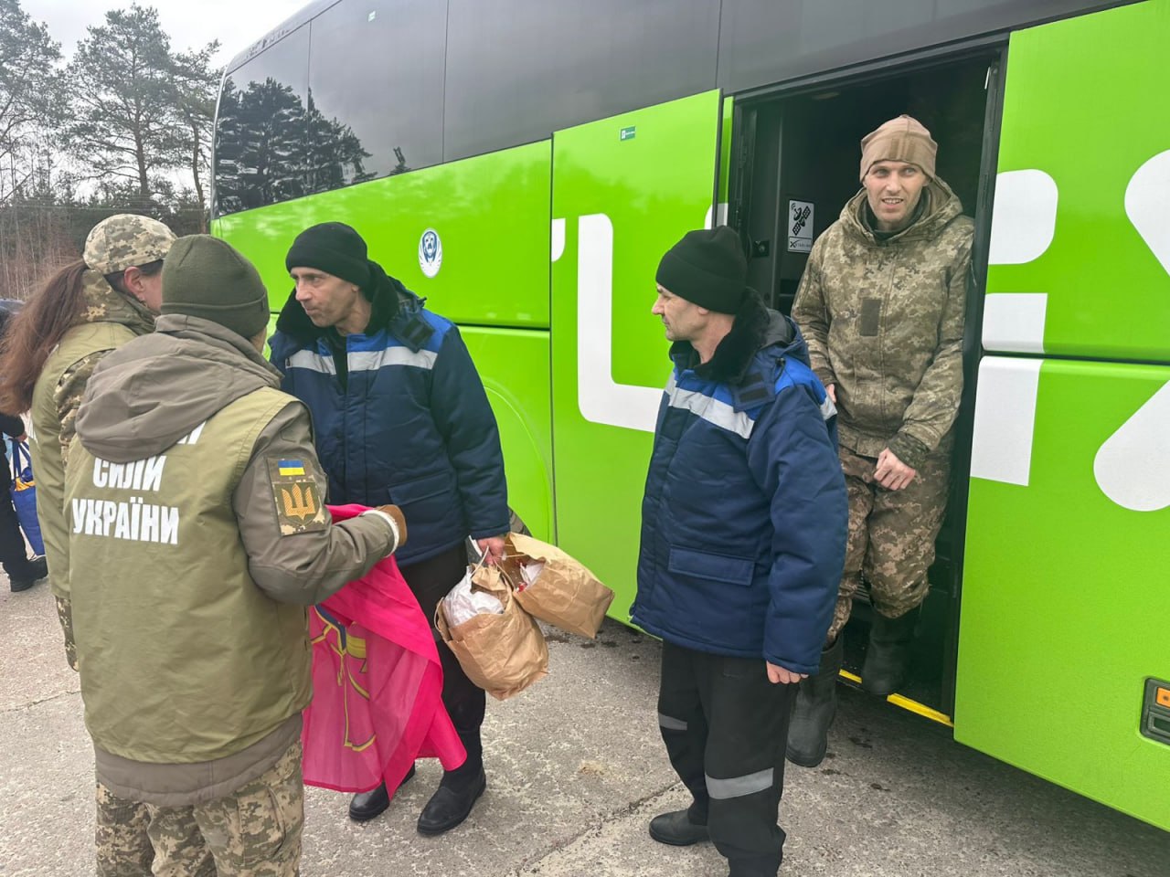 Люди у військовій формі зустрічають чоловіків, які виходять із великого зеленого автобуса. На першому плані жінка у військовій куртці з написом «Сили України» передає чоловікові паперові пакунки та рожеву тканину. Ще один чоловік у камуфляжі стоїть у дверях автобуса, спостерігаючи за процесом.