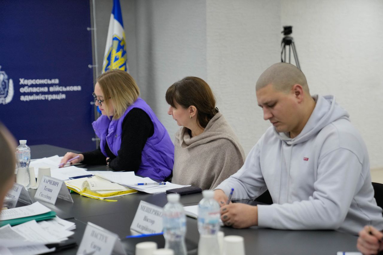 Троє людей сидять за робочим столом під час засідання, зосереджено вивчаючи документи та роблячи нотатки. На фоні — прапор України та офіційний банер «Херсонська обласна військова адміністрація».