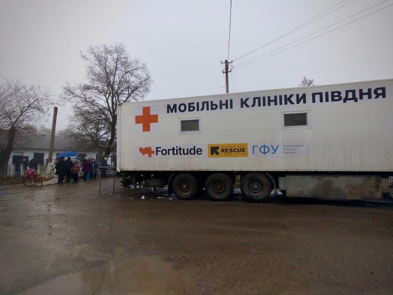 Велика біла вантажівка з написом «МОБІЛЬНІ КЛІНІКИ ПІВДНЯ» стоїть на розмитій ґрунтовій дорозі в сільській місцевості. На борту автомобіля зображено червоний хрест та логотипи організацій-партнерів. На задньому плані, біля житлових будинків, під парасольками стоїть група людей.