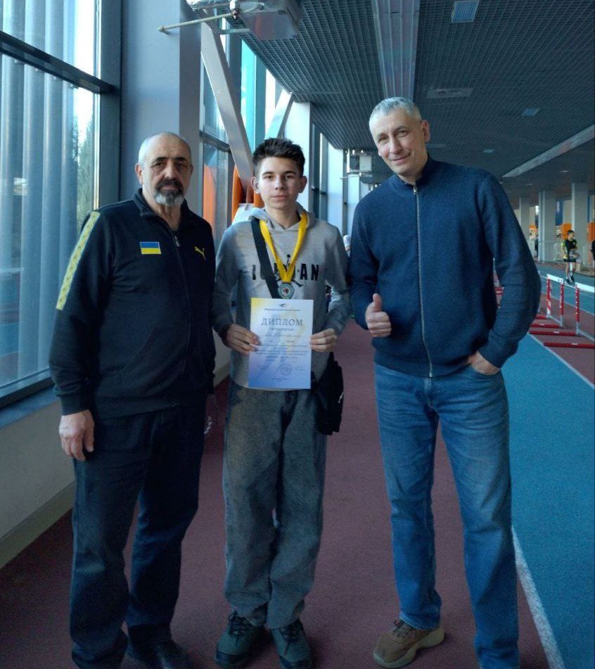 Троє осіб стоять у легкоатлетичному манежі. У центрі — юнак із медаллю на шиї, який тримає в руках диплом. Його супроводжують двоє чоловіків: один у спортивному костюмі з прапором України, інший — у синьому светрі, що показує жест «клас».