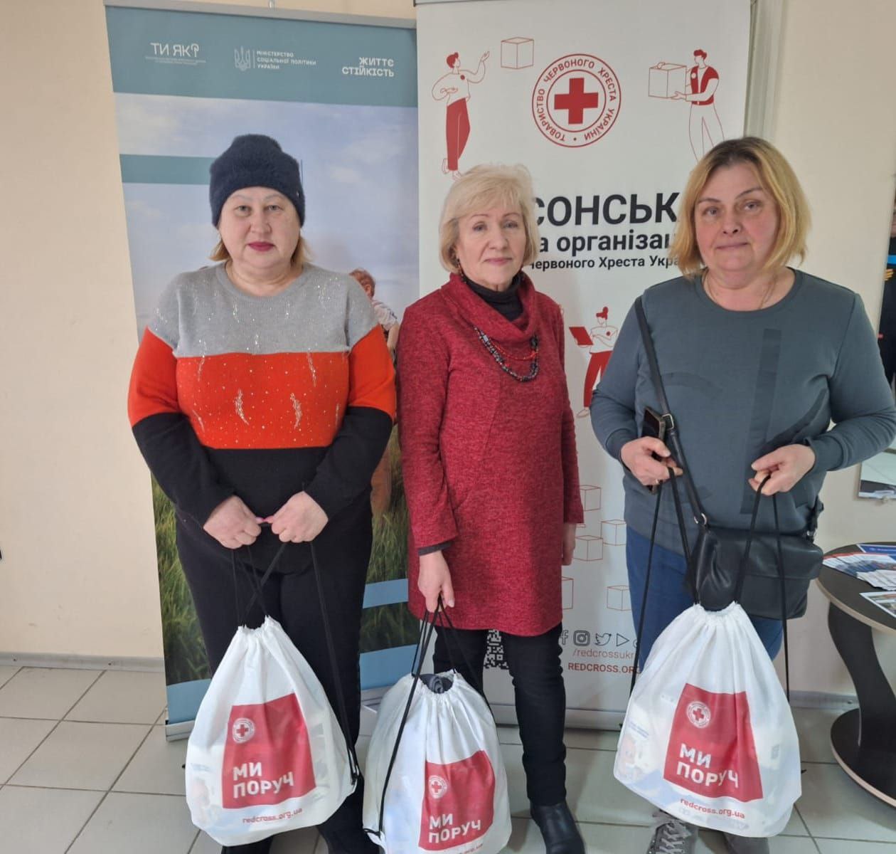 Три жінки тримають білі гуманітарні пакунки з логотипом «Ми поруч» від Товариства Червоного Хреста України. Вони стоять у приміщенні на фоні інформаційних банерів Херсонської обласної організації та програми ментального здоров'я «Ти як?». Учасниці заходу вдягнені у повсякденний теплий одяг.