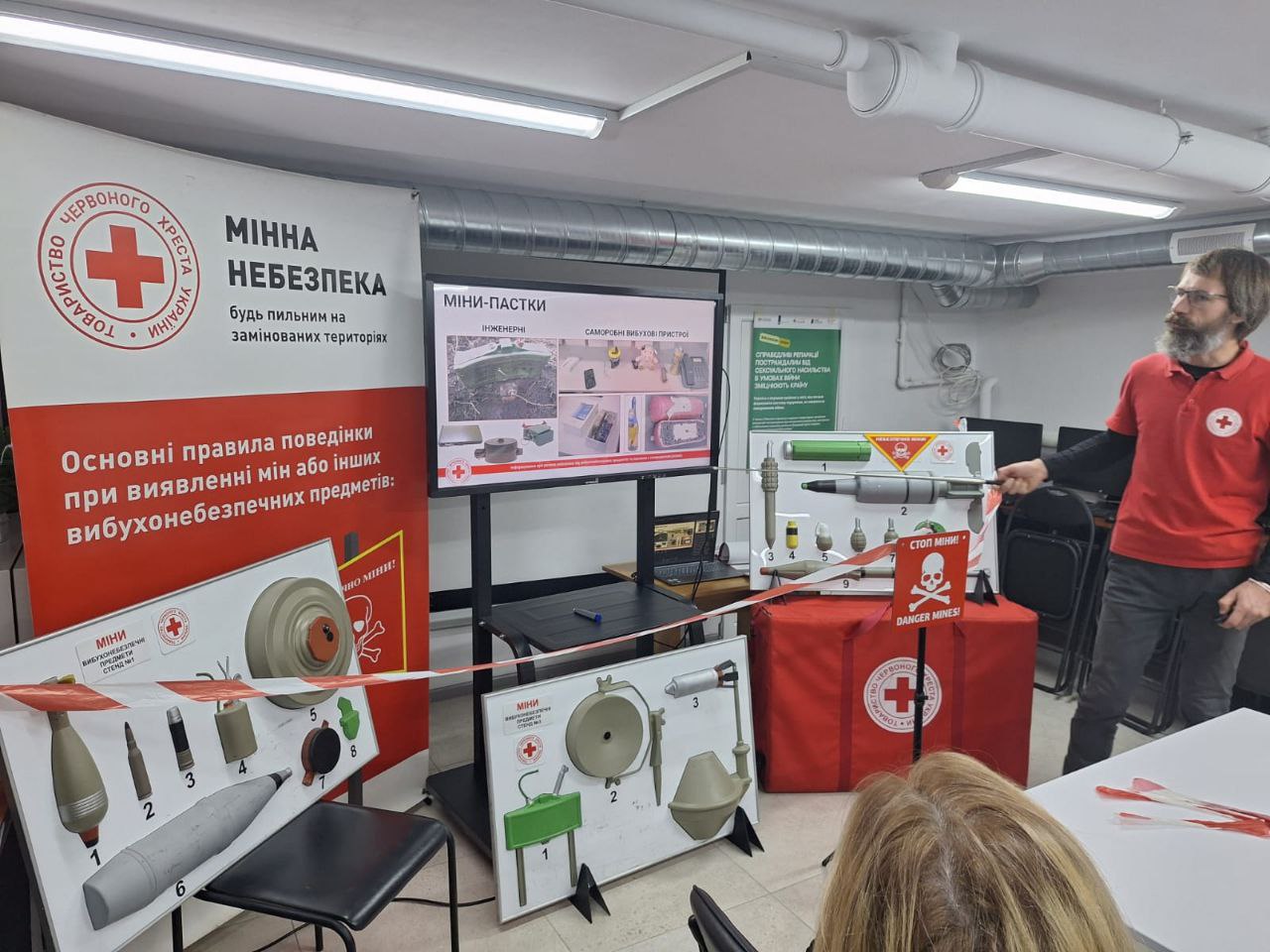 Представник Товариства Червоного Хреста України проводить інструктаж із мінної безпеки. У приміщенні розгорнуто мобільну експозицію з навчальними стендами, де представлені макети різних типів мін та боєприпасів. На екрані телевізора відображається презентація про міни-пастки та саморобні вибухові пристрої.