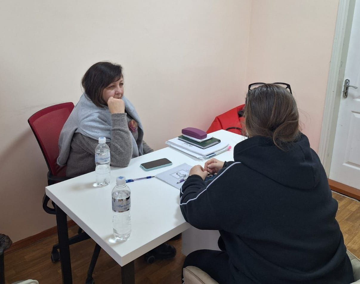 Дві жінки сидять одна навпроти одної за білим столом під час індивідуальної консультації. На столі лежать робочі документи, блокнот, смартфон та дві пляшки води. Обстановка в кабінеті нагадує зустріч із психологом або соціальним працівником у межах програми підтримки.