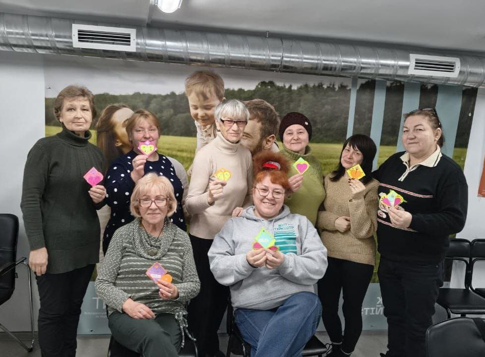 Група жінок усміхається та позує для фото, тримаючи в руках саморобні паперові обереги у формі сердечок. Вони знаходяться у приміщенні на фоні великого фотополотна із зображенням людей на природі. Атмосфера заходу свідчить про проведення майстер-класу або сесії психосоціальної підтримки.