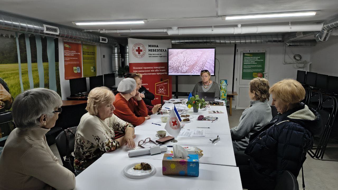 Група людей сидить за довгим столом у приміщенні, схожому на укриття, під час лекції з мінної безпеки. На фоні встановлено стенд від Червоного Хреста та екран із презентацією. Учасники слухають спікерку та переглядають інформаційні матеріали, що лежать на столі.