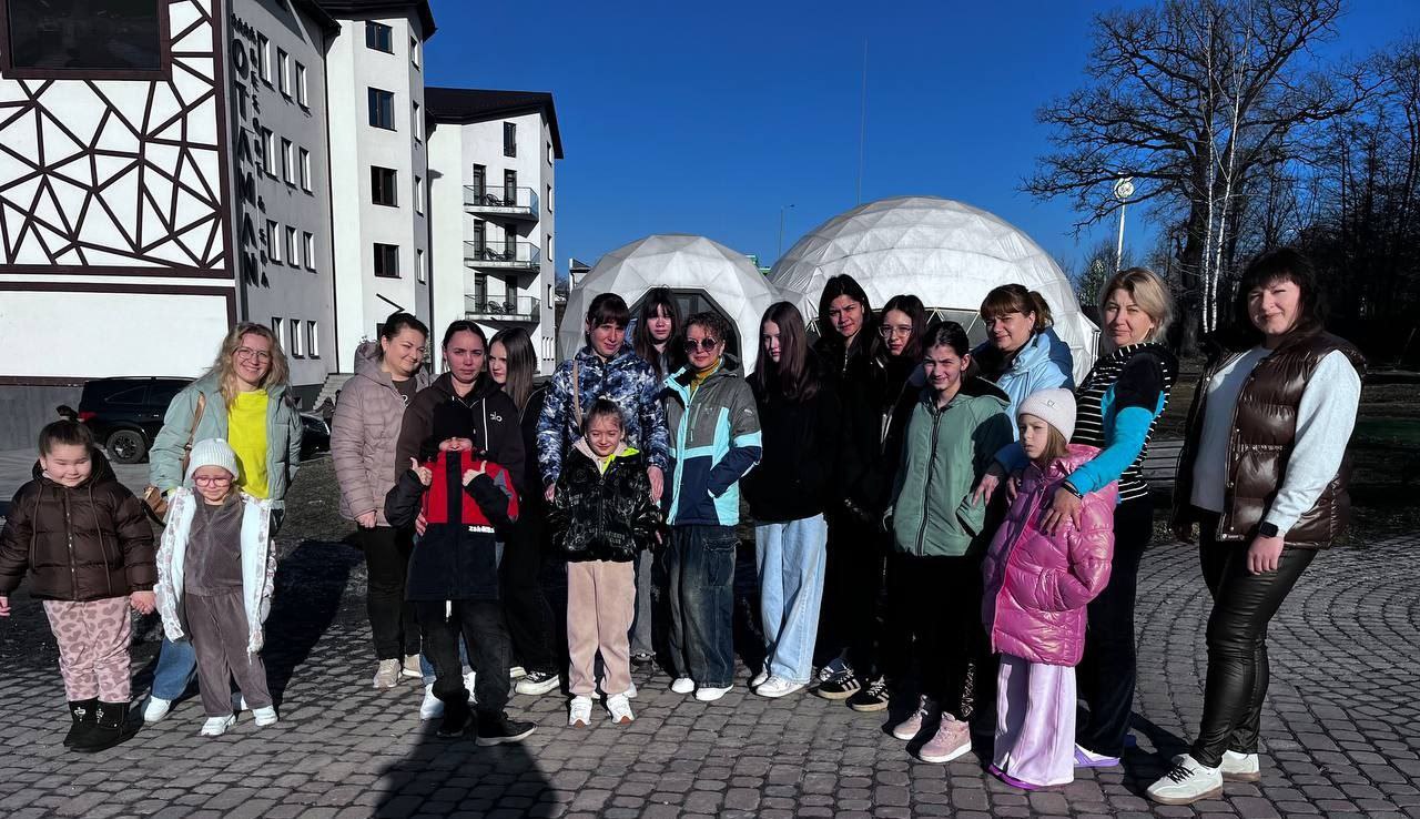 Група дітей та дорослих позує для спільного фото на фоні багатоповерхових будівель та білих купольних споруд у сонячний день.