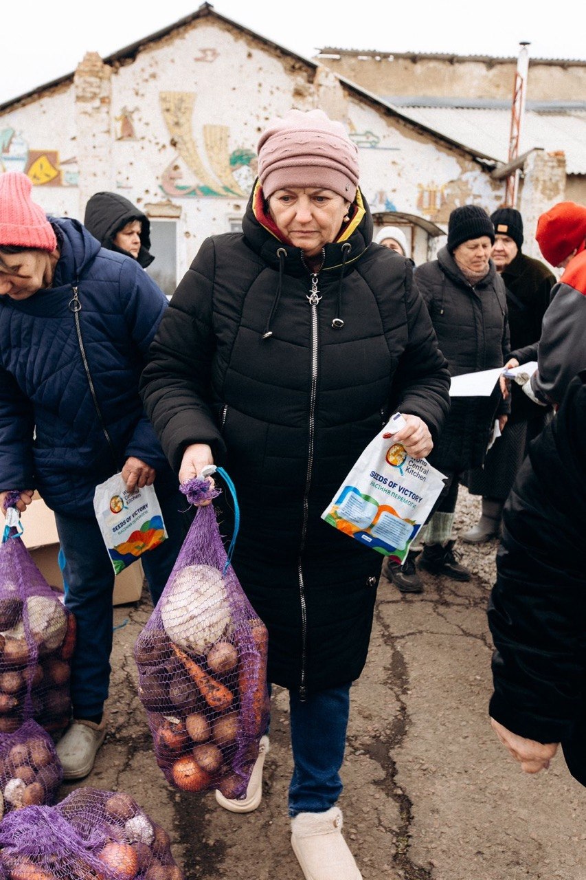 Люди отримують гуманітарну допомогу на фоні пошкодженої будівлі. У центрі жінка тримає сітку з овочами (картоплею, морквою, буряком) та пакунок насіння «Насіння перемоги» від організації World Central Kitchen.