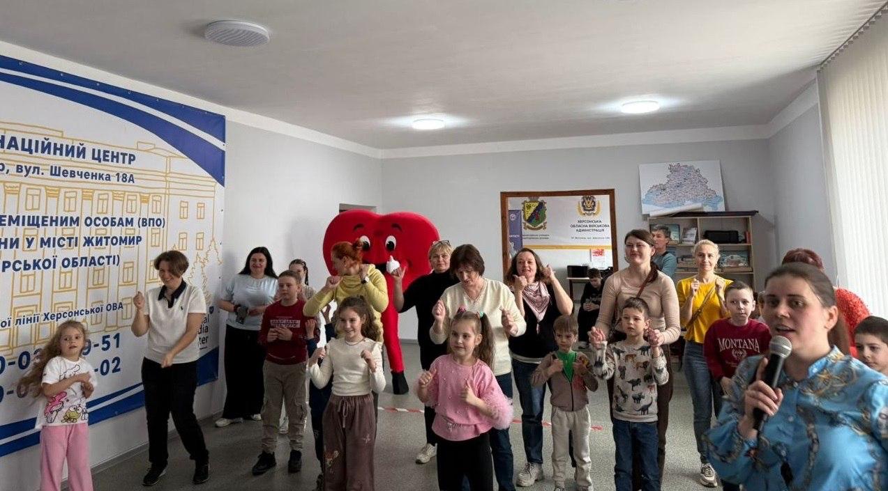 Група дорослих та дітей позує під час заходу в приміщенні Координаційного центру. Позаду стоїть аніматор у костюмі великого червоного серця, а ліворуч розміщено банер із контактами центру допомоги переселенцям із Херсонщини в Житомирі.